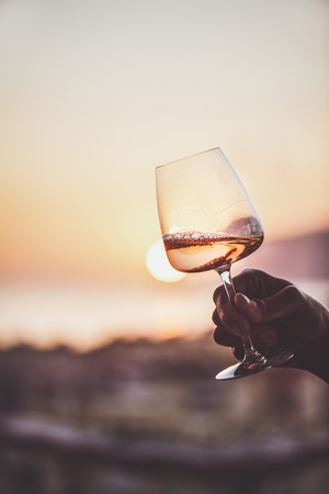 Glass of rose wine in manÂ€Â™s hand with sea view and sunset at background, close-up. Summer...