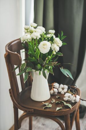 Spring white buttercup flowers in enamel jug on wooden vintage chair with white wall and curtain behind.の写真素材