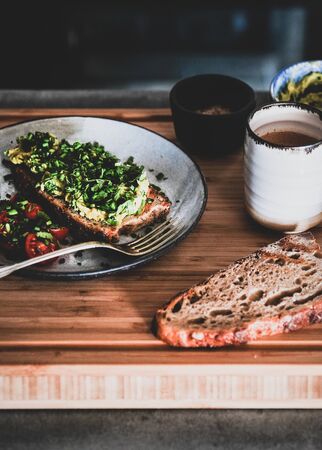 Healthy vegan summer breakfast or lunch. Avocado toast on sourdough bread with chives, coriander and cherry-tomatoes with cup of coffee on wooden board.の写真素材