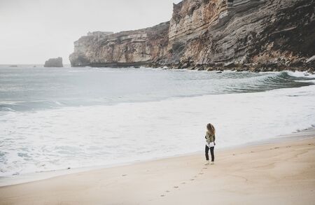 Young woman tourist walking at Atlantic ocean sandy beach in Nazare, Portugal on gloomy dayの写真素材