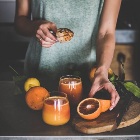 Young female making freshly squeezed blood orange juice or smoothie near concrete kitchen counter, square crop.の写真素材