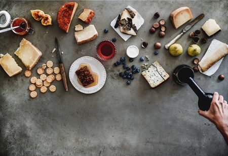Wine snack variety. Flat-lay of cheeses, honey, fruit, nuts, crackers, female hands pouring wine from bottle to glass over grey concrete background, top view, copy space. Gathering, party food conceptの写真素材