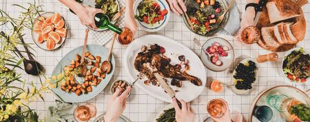 Family or friends gathering dinner. Flat-lay of hands of people eating lamb shoulder, salads, vegetables, drinking wine over checkered tablecloth, top view, wide composition. Celebration party dinnerの写真素材