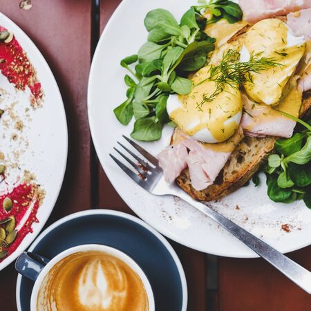 Trendy breakfast set. Flat-lay of poached Eggs Benedict on sourdough bread toast with green salad and pork ham and cup of coffee on cafe table, top view, square cropの写真素材
