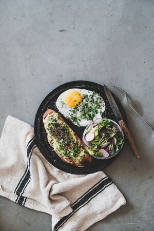 Healthy breakfast. Flat-lay of avocado toast on sourdough bread with herbs and sprouts, fried egg and salad over grey concrete background, top view, copy space. Clean eating, balanced diet conceptの写真素材