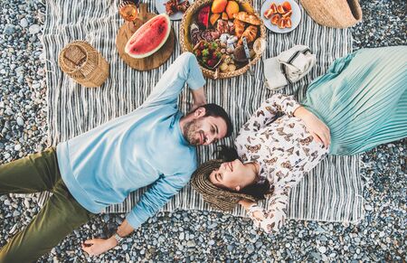 Summer beach picnic at sunset. Young couple lying on striped blanket, looking at each other at weekend picnic outdoor at seaside with sparkling wine, fresh fruit and tray of tasty appetizers, top viewの写真素材