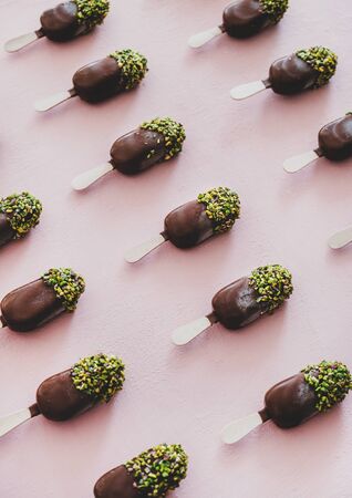 Ice-cream lolly pattern. Chocolate glazed ice-cream pops with pistachio icing in rows over pastel pink background, top view. Summer cold sweet healthy vegan dessertの写真素材