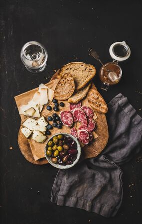 Wine and snack set. Flat-lay of grilled pieces of bread, cheese variety, salami slices, olives, apricot jam, berries and white wine over rustic black background, top viewの写真素材