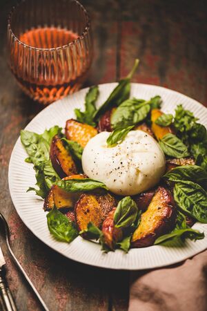 Seasonal salad with Burrata soft cheese and grilled peaches with glass of rose wine on white plate over rustic red painted table background, selective focusの写真素材