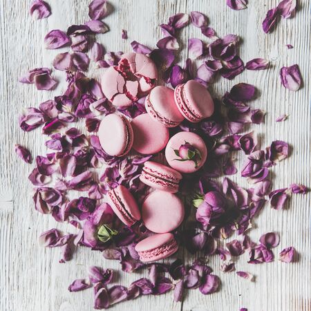 Flat-lay of sweet pink macaron cookies heap and rose buds and petals over wooden background, top view, square crop. Food texture, background and wallpaperの写真素材