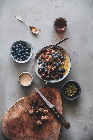 Healthy vegan breakfast. Flat-lay of quinoa oat granola coconut yogurt bowl with fresh fruit, seed, nut, berries and cup of coffee over grey background, top view. Clean eating, vegetarian food conceptの写真素材