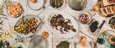 Family, friends gathering dinner. Flat-lay of roasted lamb shoulder, salads, baked vegetables, snacks, bread, rose wine and blooming mimosa branches over white tablecloth, top view, wide compositionの写真素材