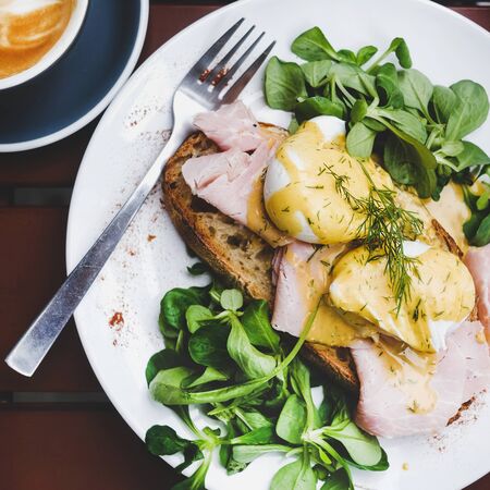 Trendy breakfast set. Flat-lay of poached Eggs Benedict on sourdough bread toast with green salad and pork ham on cafe table, top view, square cropの写真素材