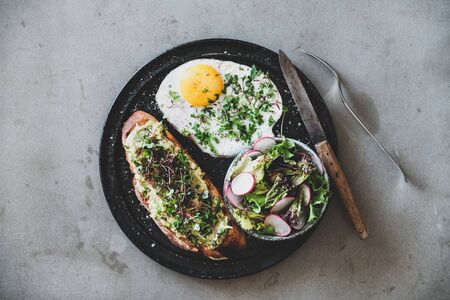 Healthy breakfast. Flat-lay of avocado toast on sourdough bread with herbs and sprouts, fried egg with coriander and salad over grey concrete background, top view. Clean eating, balanced diet conceptの写真素材