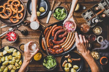 Flat-lay of Octoberfest party dinner table with grilled meat sausages, German pretzel pastry, potatoes, cucumber salad, sauces, beers and peoples hands with food over dark wooden background, top viewの写真素材