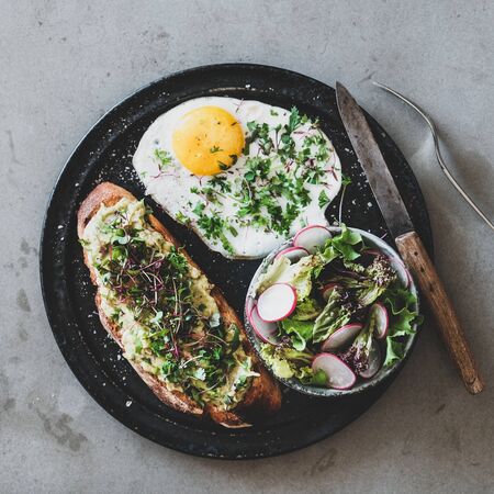 Healthy breakfast. Flat-lay of avocado toast on sourdough bread with herbs and sprouts, fried egg with coriander and salad over grey concrete background, top view, square crop. Clean eating conceptの写真素材