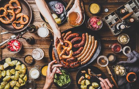 Flat-lay of Octoberfest party dinner table with grilled meat sausages, pretzel pastry, potatoes, cucumber salad, sauces, beers and peoples hands holding food over dark wooden background, top viewの写真素材