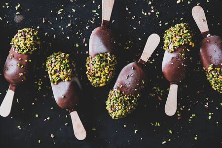 Ice cream pattern. Flat-lay of chocolate glazed ice cream pops with pistachio icing over dark background, top view, close-up. Summer seasonal cold sweet healthy vegan dessertの写真素材