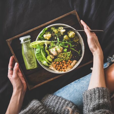 Healthy dinner, lunch in bed. Vegan superbowl or Buddha bowl with hummus, vegetable, fresh salad, beans, couscous and avocado, green smoothie on tray and woman in jeans eating, top view, square cropの写真素材
