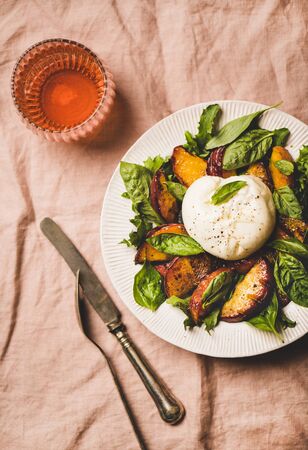 Flat-lay of seasonal salad with Burrata soft cheese and grilled peaches with glass of rose wine on white plate over pink pastel linen tablecloth, top viewの写真素材