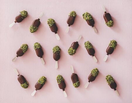 Ice-cream pattern. Flat-lay of chocolate glazed ice-cream pops with pistachio nuts icing over pastel pink background, top view. Summer cold sweet healthy vegan dessertの写真素材
