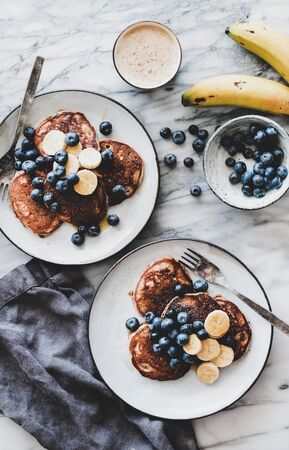 Healthy comfort breakfast set. Flat-lay of banana pancakes with blueberry and honey and cup of coffee over white marble table background, top viewの写真素材