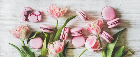 Flat-lay of sweet pink macaron cookies and fresh spring tulip flowers over white wooden background, top view, wide composition. Food texture, background and wallpaperの写真素材
