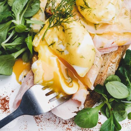 Trendy breakfast. Flat-lay of poached Eggs Benedict on sourdough bread toast with green salad and pork ham on white plate, close-up, top view, square cropの写真素材