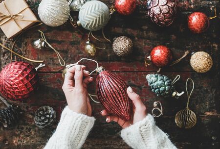 Christmas or New Year holiday background, texture, wallpaper. Flat-lay of Christmas festive decoration toys and wonans hands holding red toy over red rustic wooden background, top viewの写真素材