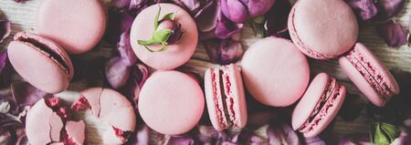 Flat-lay of sweet pink macaron cookies and rose buds and petals over wooden background, top view, close-up, wide composition. Food texture, background and wallpaperの写真素材