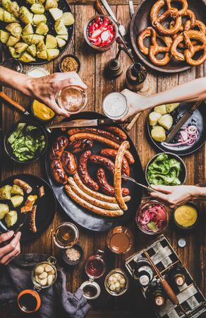 Flat-lay of Octoberfest party dinner table with grilled sausages, pretzel pastry, potatoes, cucumber salad, sauces, beers and peoples hands with food and glasses over dark wooden background, top viewの写真素材