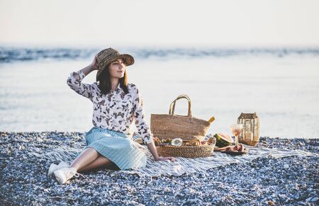 Summer beach picnic at sunset. Young woman in hat sitting on blanket and having weekend picnic outdoor at seaside with fresh seasonal fruit and tray full of of tasty appetizers and looking awayの写真素材