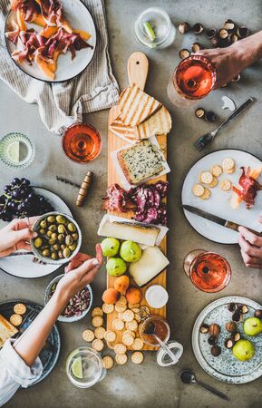 Mid-summer picnic with wine and snacks. Flat-lay of charcuterie and cheese board, rose wine, nuts, olives and peoples hands holding glasses and food over concrete table background, top viewの写真素材