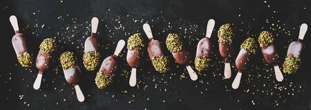 Ice cream pattern. Flat-lay of chocolate glazed ice cream pops with pistachio icing over dark background, top view, wide composition. Summer seasonal cold sweet healthy vegan dessertの写真素材