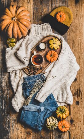 Autumn outfit layout. Flat-lay of beige knitted sweater, blue jeans, hat, decorative pumpkins, candle and tea in cup over rustic wooden background, top view. Fall apparel and moodの写真素材