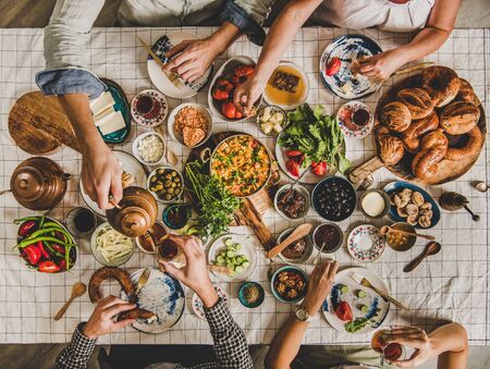 Flat-lay of people sitting at table with Turkish breakfast with pastries, vegetables, greens, cheeses, eggs, jams and tea in tulip glasses and copper teapots over chekered linen tablecloth, top viewの写真素材