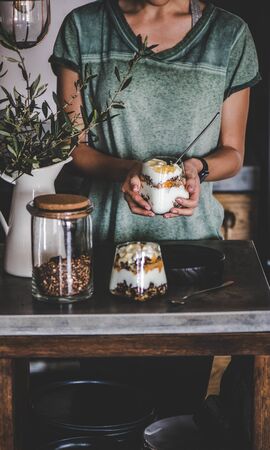 Healthy vegan breakfast. Woman holding glass with greek or coconut yogurt with granola and mango smoothie layers with fruit in hands.の写真素材
