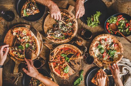 Family or friends having pizza party dinner. Flat-lay of people eating various kinds of Italian pizza and drinking wine over wooden table, top view. Fast food lunch, gathering, celebration conceptの写真素材