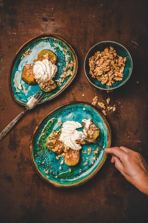 Turkish traditional pumpkin dessert. Flat-lay of pumpkin in sweet syrup with cinnamon, walnuts, whipped cream and human hand with fork, top view. Middle East oriental cuisine typical vegan foodの写真素材