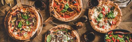Pizza party dinner. Flat-lay of various kinds of Italian pizza, salad and red wine in glasses over rustic wooden table, top view, wide composition. Fast food lunch, celebration, gathering conceptの写真素材