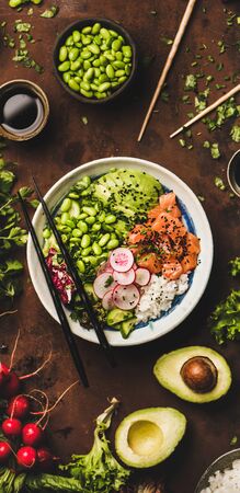 Healthy lunch. Flat-lay of salmon poke bowl or sushi bowl with various vegetables, greens, sushi rice, soy sauce over rusty table background, top view, narrow composition. Traditional Hawaiian foodの写真素材