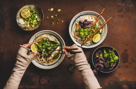 Asian cuisine lunch. Flat-lay of Vietnamese rice noodle chicken soup Pho Ga with cilantro, soy sprouts, greens in bowl and woman hands with chopsticks and lime over dark rusty background, top viewの写真素材