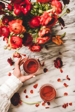 Flat-lay of womans hand holding glass of deep rose wine and bouquet of red ranunculus flowers over white marble table background, top view. Rose wine season conceptの写真素材