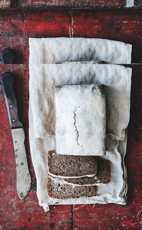 Flat-lay of healthy rye Swedish bread loaf cut in slices with vintage knife on white towel over dark red woodenの写真素材