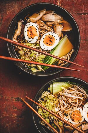 Flat-lay of traditional japanese Ramen soup with chicken meat and shiitake mushrooms in black bowls and bamboo chopsticks over dark red wooden table background, top view. Japanese cuisine conceptの写真素材