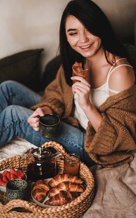 Young beautiful smiling brunette woman in comfortable casual clothing enjoying breakfast in bedの写真素材