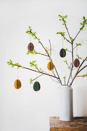 Easter holiday home decoration. Tree branches in vase with fresh spring leaves decorated with festive colorful Easter eggs, white wall background. Easter holiday preparation, greeting cardの写真素材
