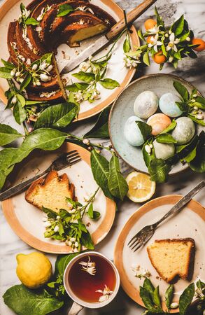 Easter holiday dinner table setting. Flat-lay of colorful Easter Eggs, Lemon cake with citrus tree blossom branches and tea over white marble table background, top view. Easter greeting cardの写真素材
