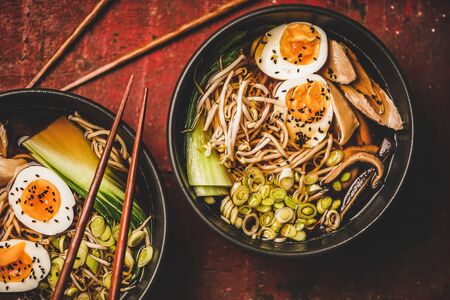 Flat-lay of traditional japanese Ramen soup with chicken meat, eggs, shiitake mushrooms in black bowls and bamboo chopsticks over dark red wooden table background, top view. Japanese cuisine conceptの写真素材