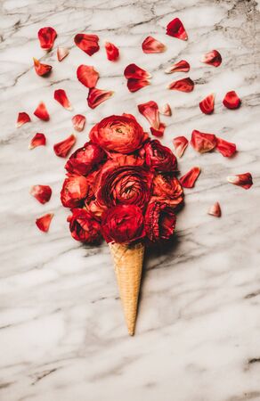 Flat-lay of ice cream waffle cone with scoop of red blooming ranunculus flowers over white marble table with petals.の写真素材